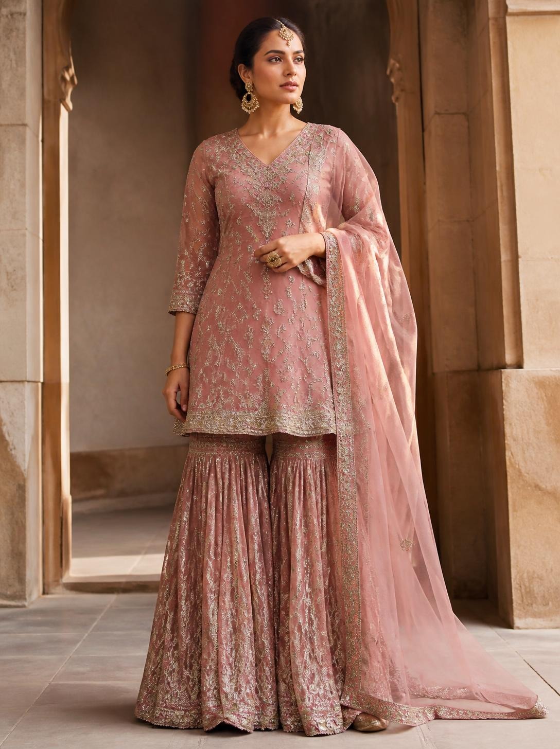Woman in a dusty-rose embroidered kurta and sharara with a sheer dupatta in a sandstone archway