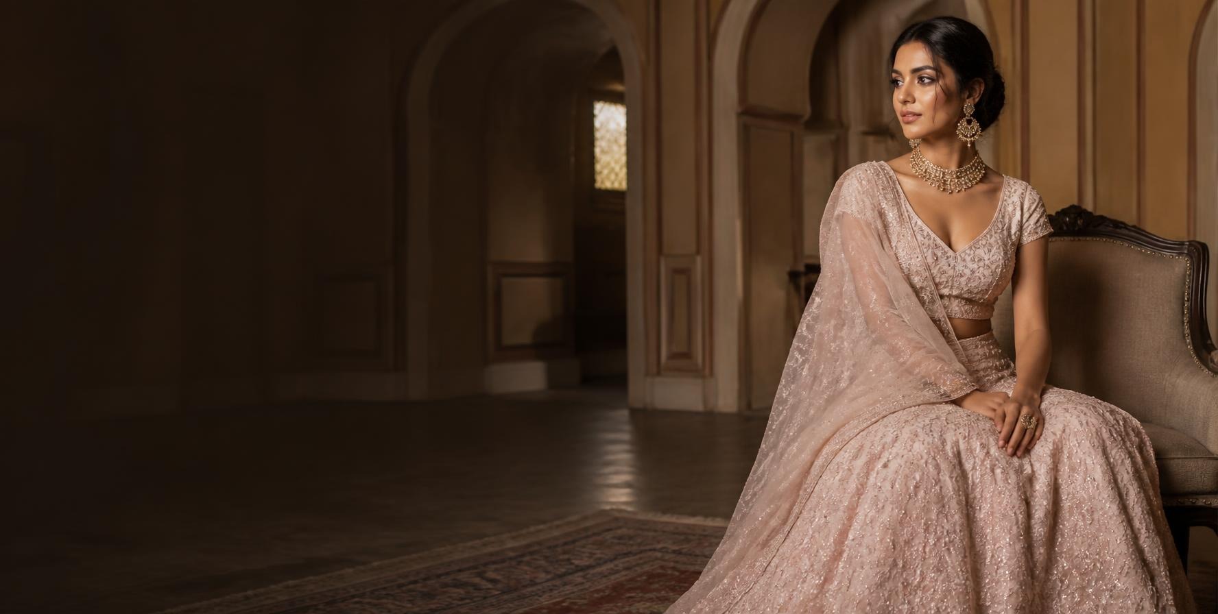 Woman in a blush embroidered lehenga with a sheer dupatta seated on an antique chair in a heritage hall